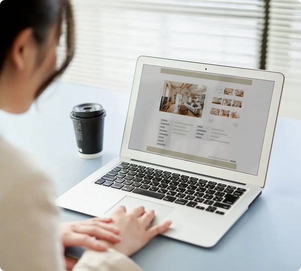 Donna di spalle consulta su un laptop argentato un sito con foto di interni, accanto un bicchiere da caffè nero