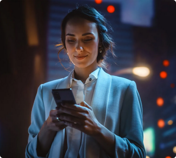 Donna in giacca azzurra e camicia bianca sorride guardando lo smartphone, di sera tra luci sfocate della città